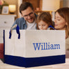 A family of three - a man, a woman, and a young girl - are gathered around a large white and blue storage bag with the name "William" printed on it.