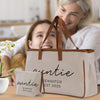 A woman and a young girl are smiling and embracing each other in a kitchen setting, with a large beige tote bag with the name "Auntie" printed on it in the foreground.