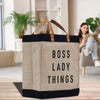 A large tote bag with the text "BOSS LADY THINGS" printed on it, placed on a wooden surface. In the background, there is a person sitting on a couch and reading a book.