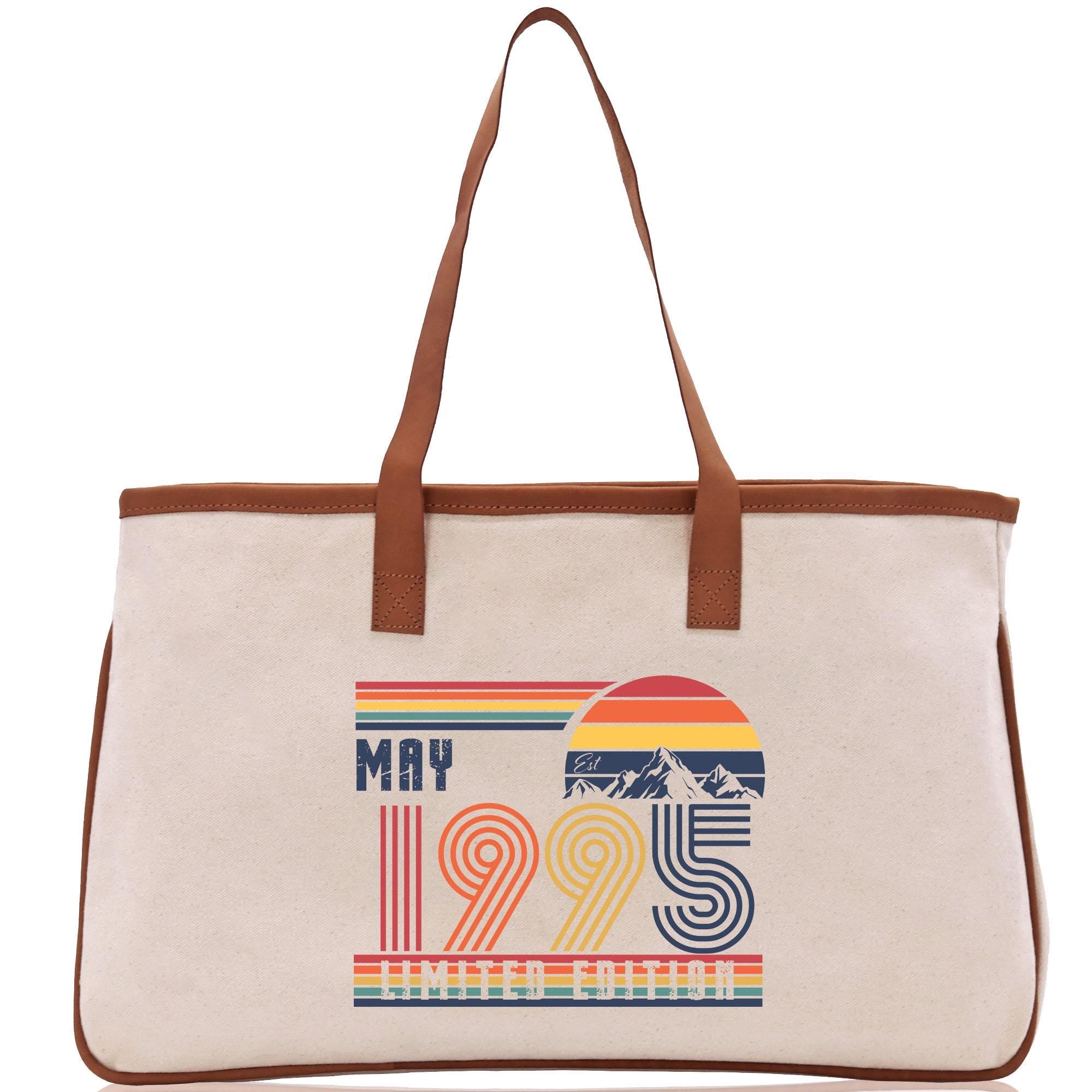 A beige canvas tote bag with a printed design on the front. The design features the text "MAY 1995" and a graphic of a sunset with mountains.