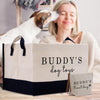 A woman is sitting on a couch, smiling and petting a dog, while a large bag labeled "Buddy's dog toys" is placed in front of her.