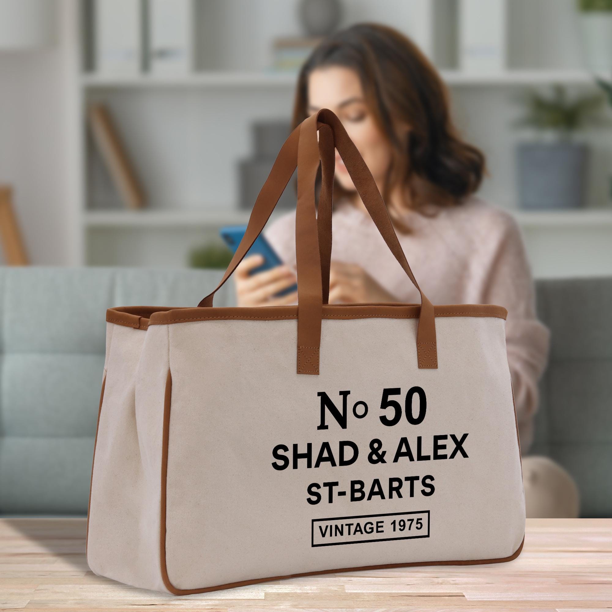 A beige canvas tote bag with the text &quot;No 50 SHAD & ALEX ST-BARTS VINTAGE 1975&quot; printed on it, sitting on a wooden surface.