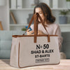 A beige canvas tote bag with the text "No 50 SHAD & ALEX ST-BARTS VINTAGE 1975" printed on it, sitting on a wooden surface.