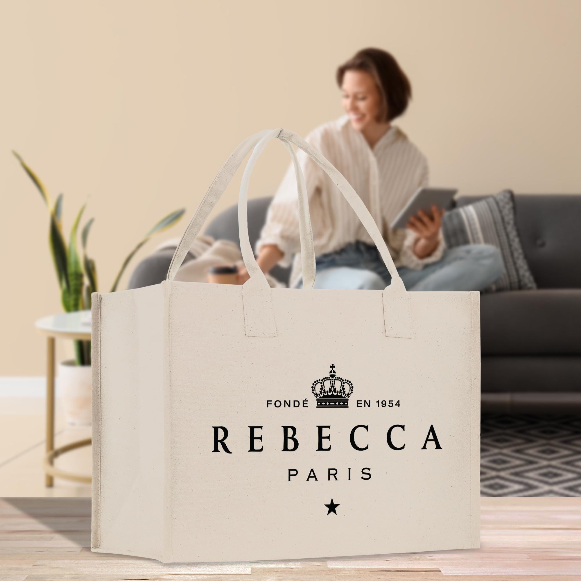 a large white tote bag with text and a crown design, placed on a wooden floor in a room with a woman sitting on a couch in the background.