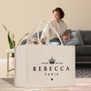 a large white tote bag with text and a crown design, placed on a wooden floor in a room with a woman sitting on a couch in the background.