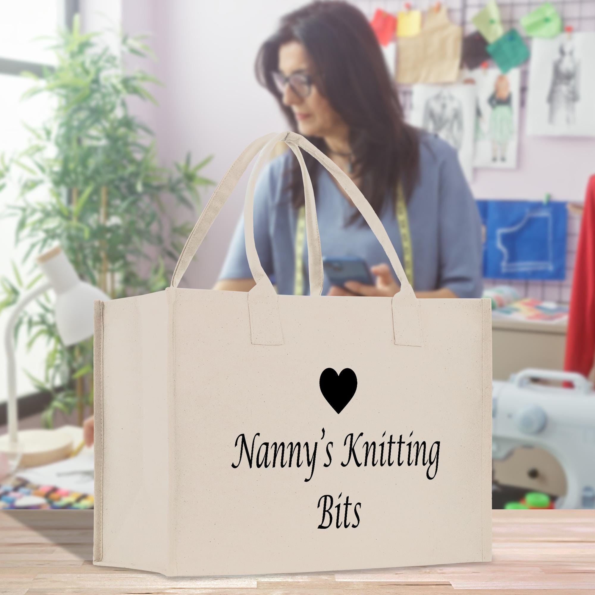 A white tote bag with the text &quot;Nanny&#39;s Knitting Bits&quot; printed on it, placed on a wooden surface. In the background, there is a woman who appears to be focused on something, possibly working on a craft project.
