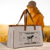 A beige canvas tote bag with a vintage-style graphic of a cow and the text "Grassmeere Cattle Ranch EST. 1950" printed on it, placed on a wooden surface. In the background, there is a blurred image of a person wearing a hat and glasses.