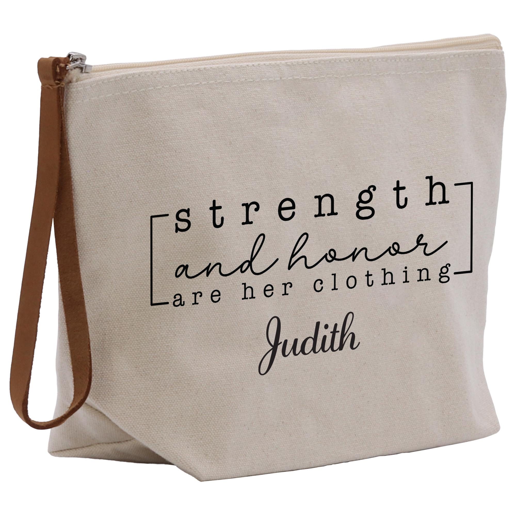 The image is a beige canvas tote bag with a zipper closure and a leather strap. It has a printed message on it that says &quot;strengths and honor are her clothing&quot; and the name &quot;Judith&quot; below it.