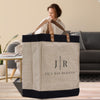A large beige tote bag with black trim and leather handles, featuring the text "JIL'L RAE DESIGNS" printed on it, is placed on a wooden surface. In the background, there is a person sitting on a couch.