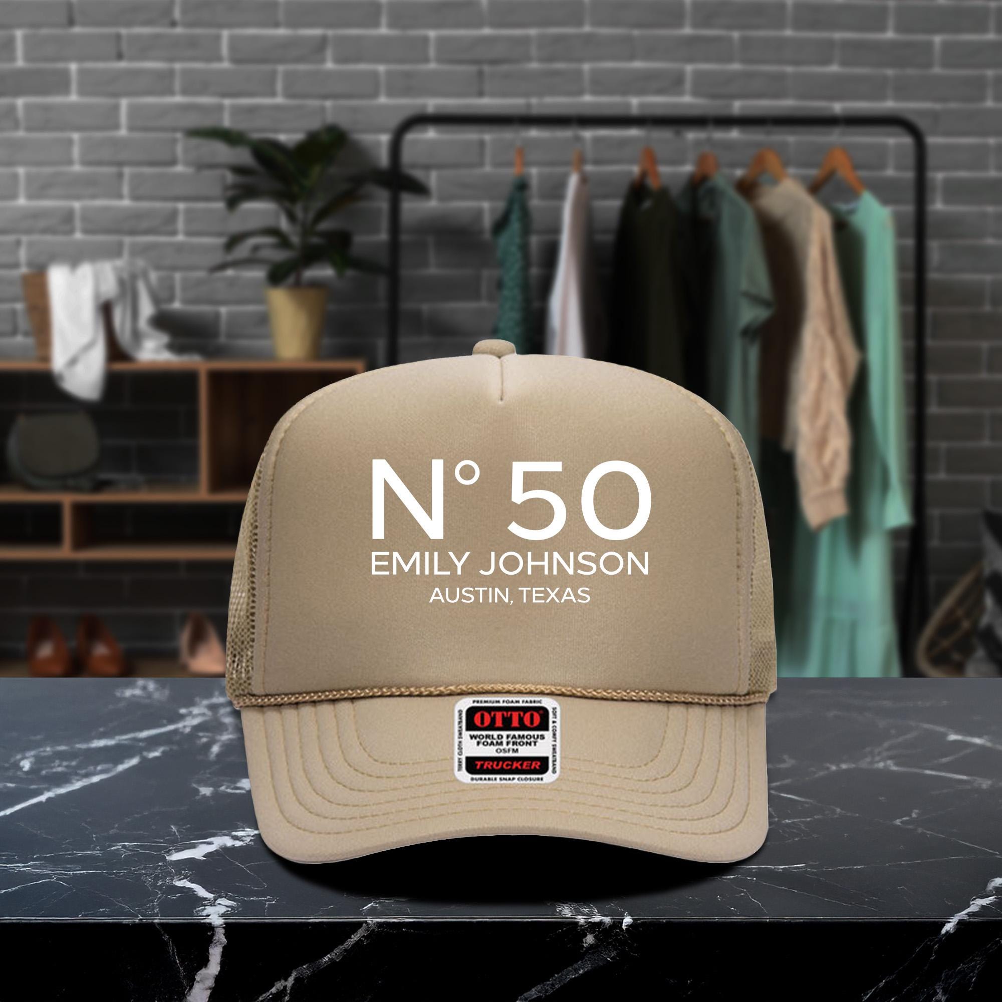A beige baseball cap with the text &quot;N° 50 EMILY JOHNSON AUSTIN, TEXAS&quot; printed on it, placed on a marble countertop.