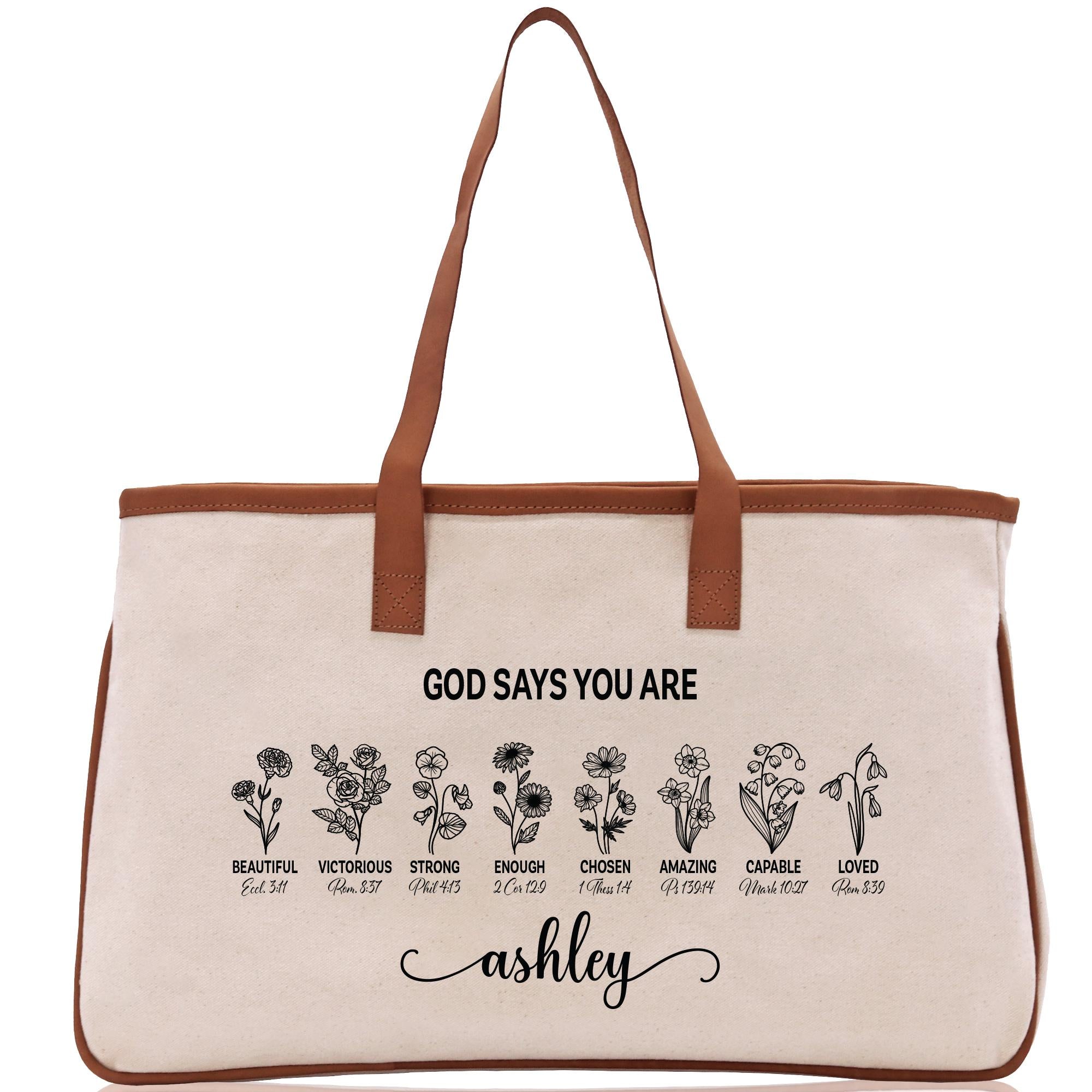 A beige canvas tote bag with a brown leather strap and the text &quot;GOD SAYS YOU ARE&quot; printed on it. Below the text, there are illustrations of flowers with descriptions of the corresponding Bible verse titles.