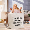 A woman sitting on a couch in a living room, holding a phone, with a white bag with text on it in front of her.