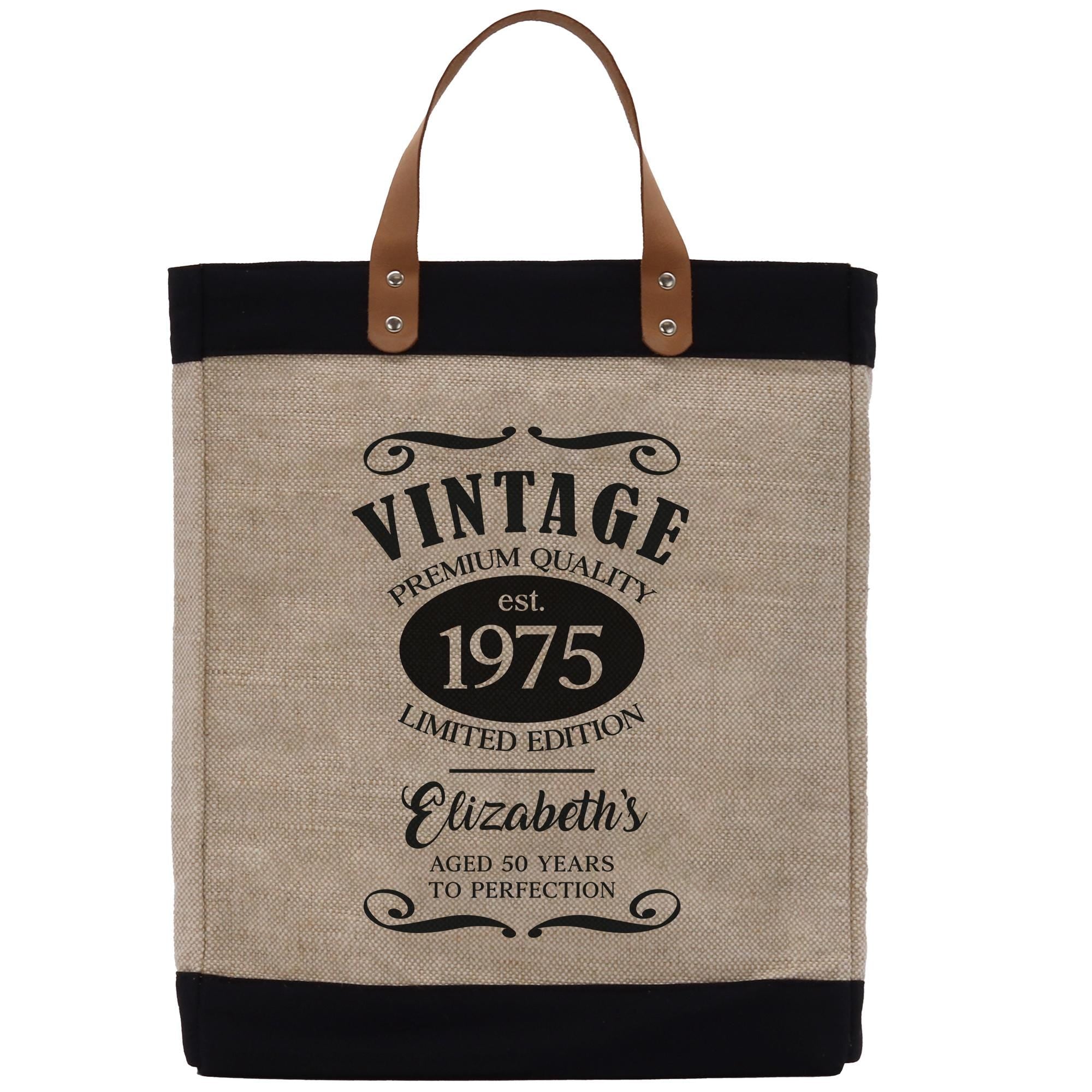 A beige tote bag with a black handle and strap, featuring a vintage-style design with text and decorative elements.