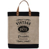A beige tote bag with a black handle and strap, featuring a vintage-style design with text and decorative elements.