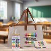 A beige tote bag with a straw and a small container labeled "Miss Magi" are displayed on a wooden surface, with a blurred classroom setting in the background.