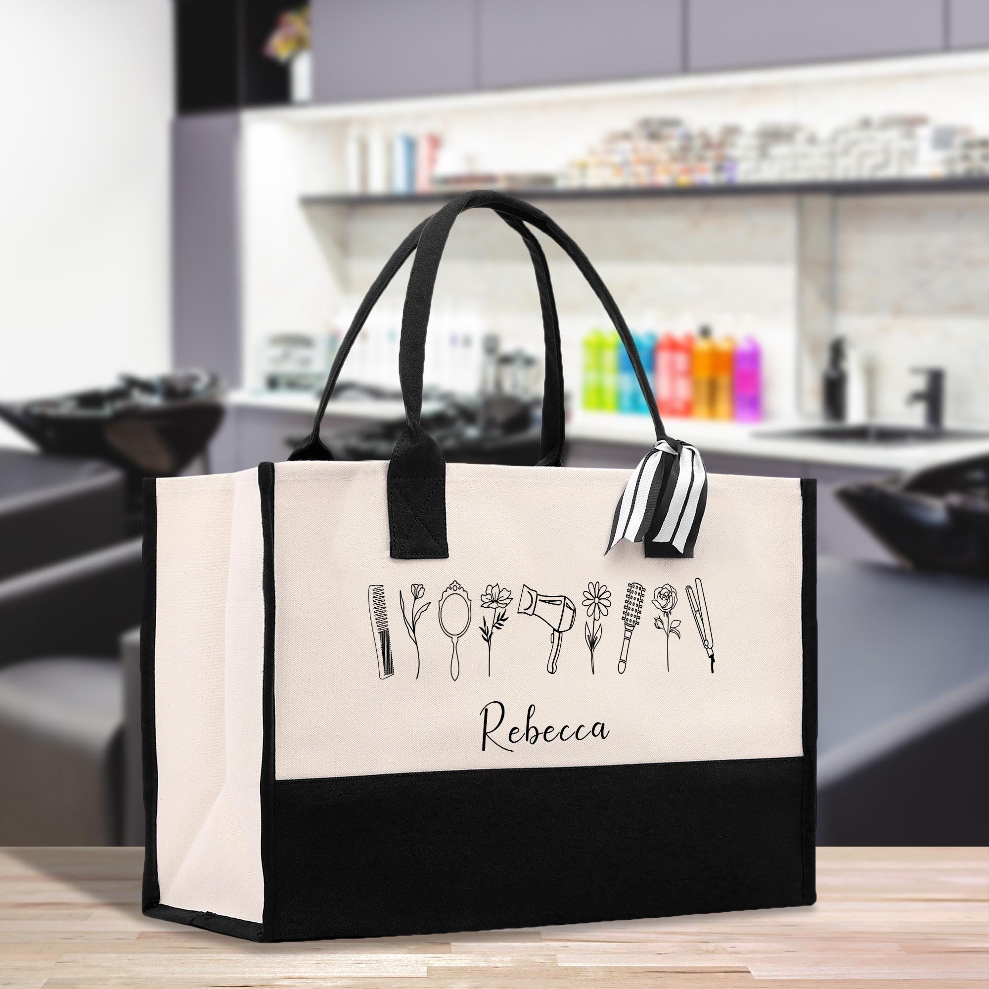 A large tote bag with a black and white design featuring various hair styling tools, such as scissors, combs, and hair dryers, printed on it. The bag has a black handle and a white trim.