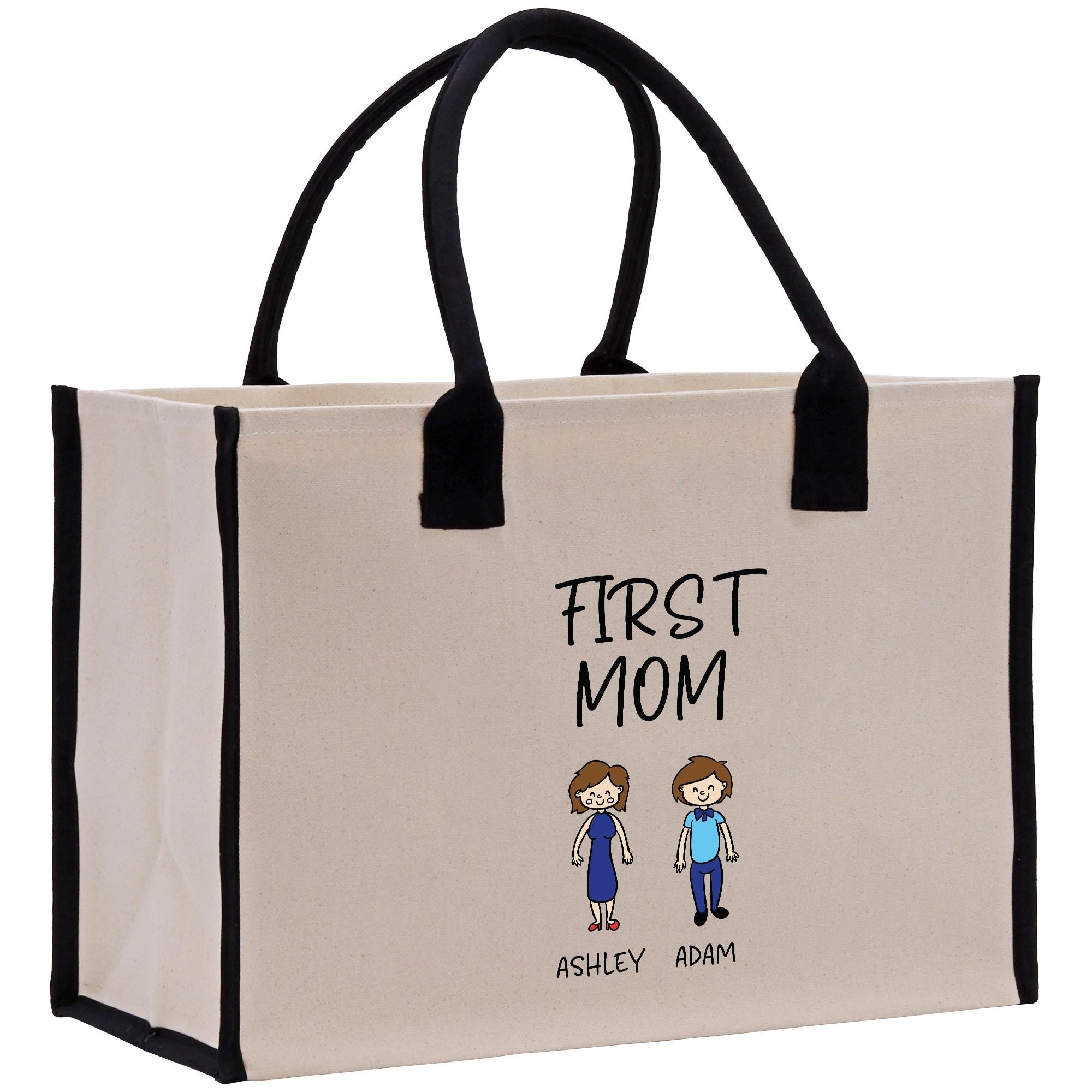 A beige canvas tote bag with black handles and straps, featuring a cartoon illustration of two people and the text &quot;FIRST MOM&quot; and &quot;ASHLEY ADAM&quot; printed on it.