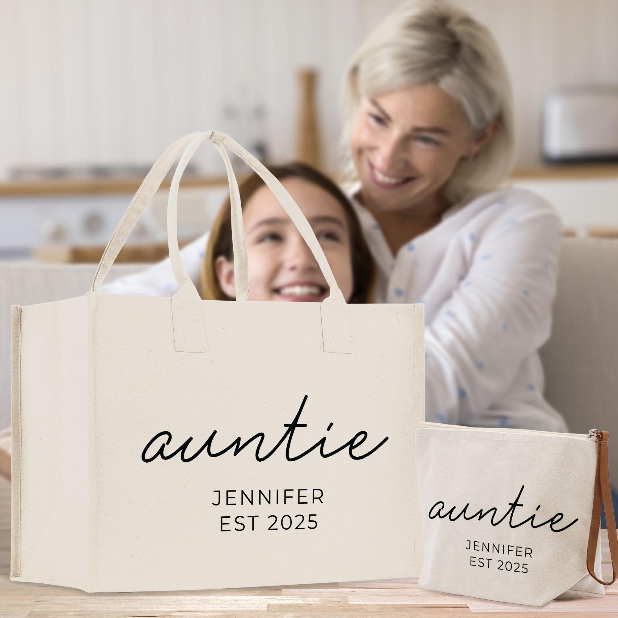 A smiling woman and a young girl are sitting together in a kitchen, with a large tote bag in the foreground that says &quot;auntie&quot; and &quot;JENNY EST 2025&quot;.