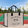 A beige tote bag with black handles and straps, featuring a tennis club logo and text, placed on a wooden surface against a backdrop of a tennis court and trees.