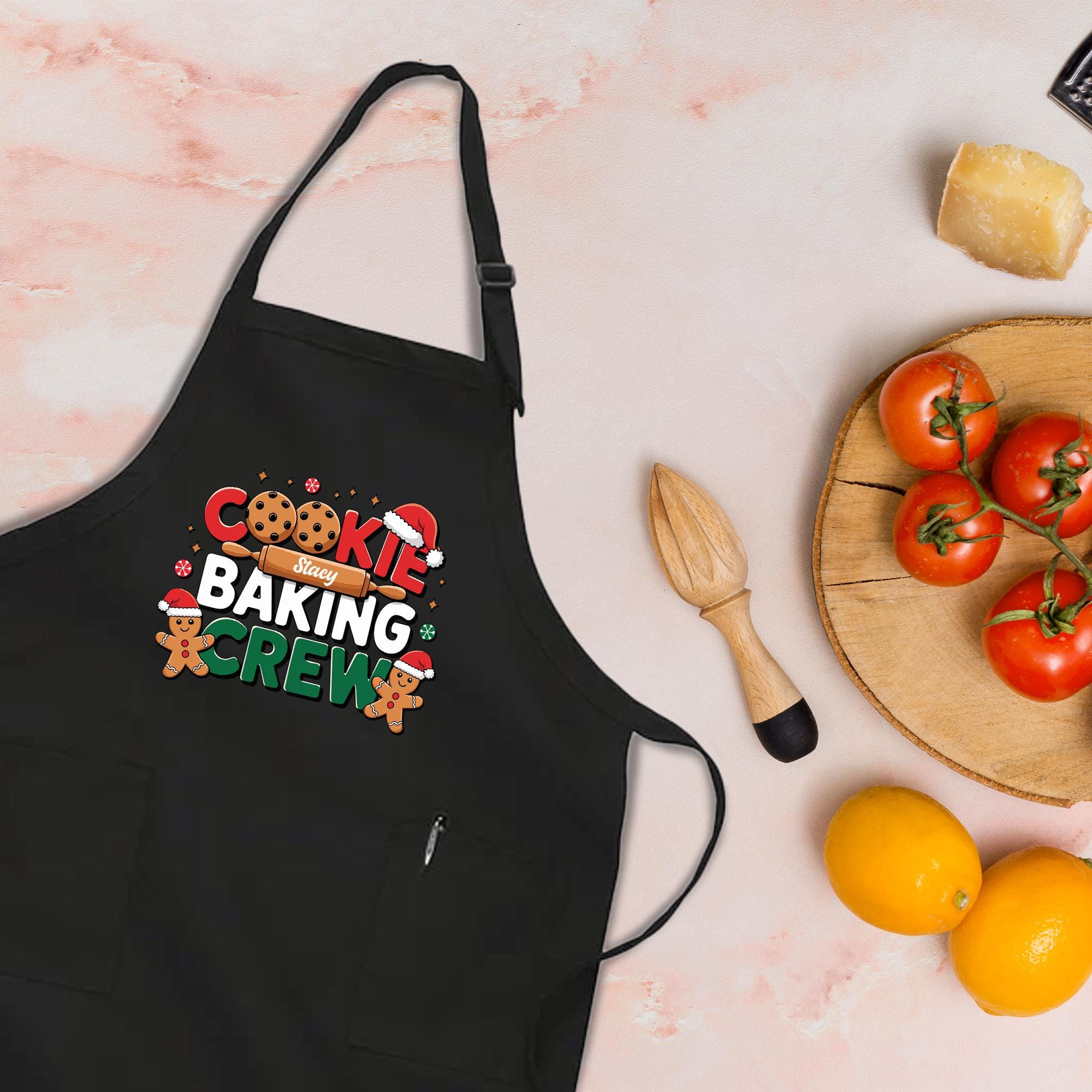 A black apron with a cookie baking crew design, a wooden juicer, a plate of tomatoes, and a lemon on a table.
