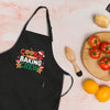 A black apron with a cookie baking crew design, a wooden juicer, a plate of tomatoes, and a lemon on a table.
