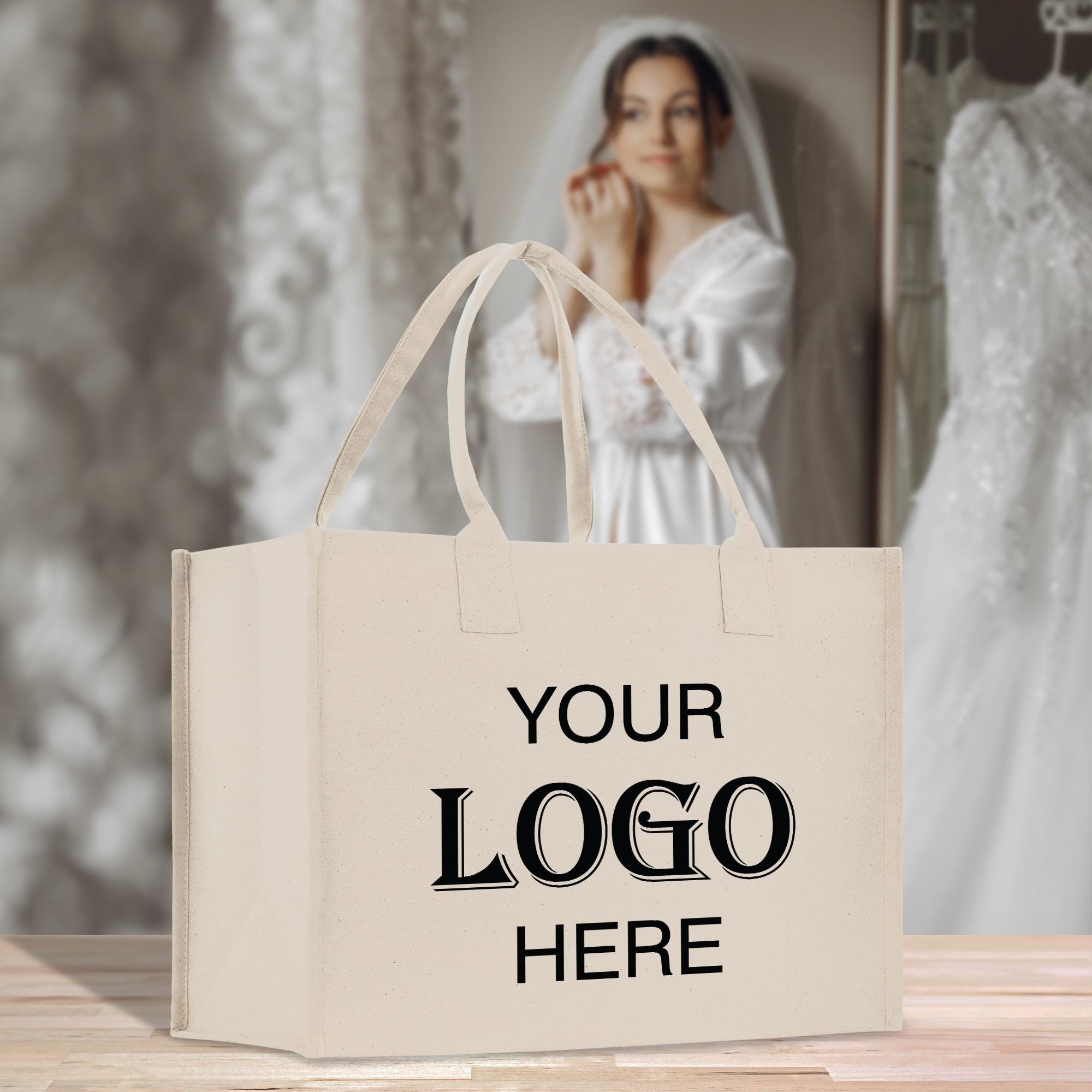 A beige tote bag with the text &quot;YOUR LOGO HERE&quot; printed on it, placed on a wooden surface. In the background, there is a blurred image of a woman wearing a white wedding dress.