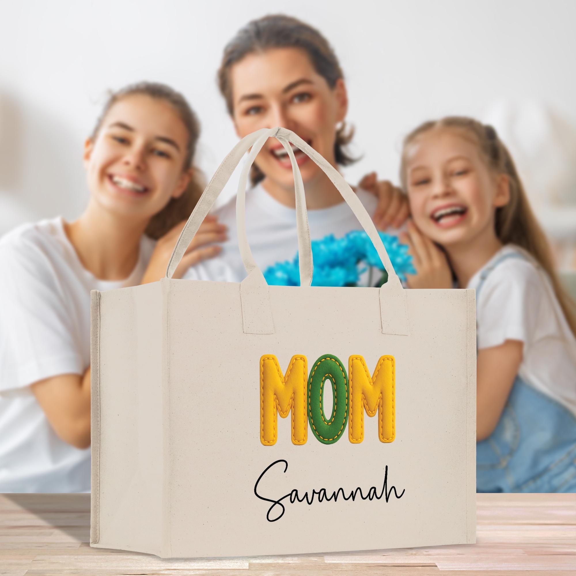 A white tote bag with the word &quot;MOM&quot; printed on it, along with the name &quot;Savannah&quot; written below it. The bag is placed in front of three smiling women, who appear to be a mother and her two daughters.