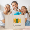 A white tote bag with the word "MOM" printed on it, along with the name "Savannah" written below it. The bag is placed in front of three smiling women, who appear to be a mother and her two daughters.