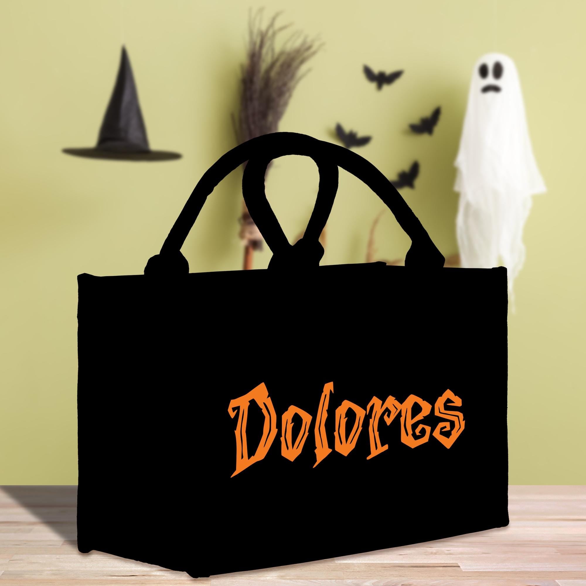 A black tote bag with the word &quot;Dolores&quot; printed on it, placed on a wooden surface. In the background, there is a witch&#39;s hat, a broom, and a ghost.