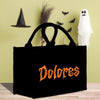 A black tote bag with the word "Dolores" printed on it, placed on a wooden surface. In the background, there is a witch's hat, a broom, and a ghost.