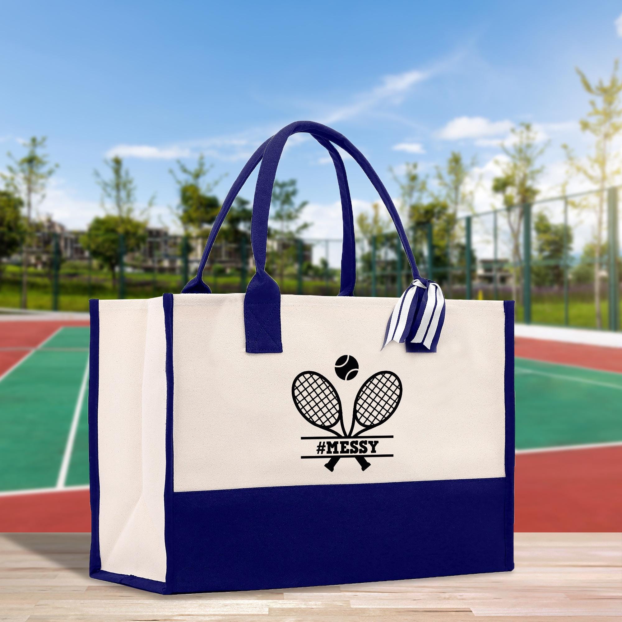 A white and blue tote bag with a tennis racket and ball design printed on it, placed on a wooden surface against a blurred background of a tennis court.