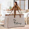 A beige canvas tote bag with a floral design and the word "Manny" printed on it, placed on a wooden surface in a living room setting.
