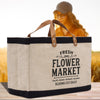 A large canvas tote bag with the text "FRESH FLOWER MARKET" printed on it, sitting on a wooden surface. In the background, there is a blurred image of a person wearing a hat and glasses.