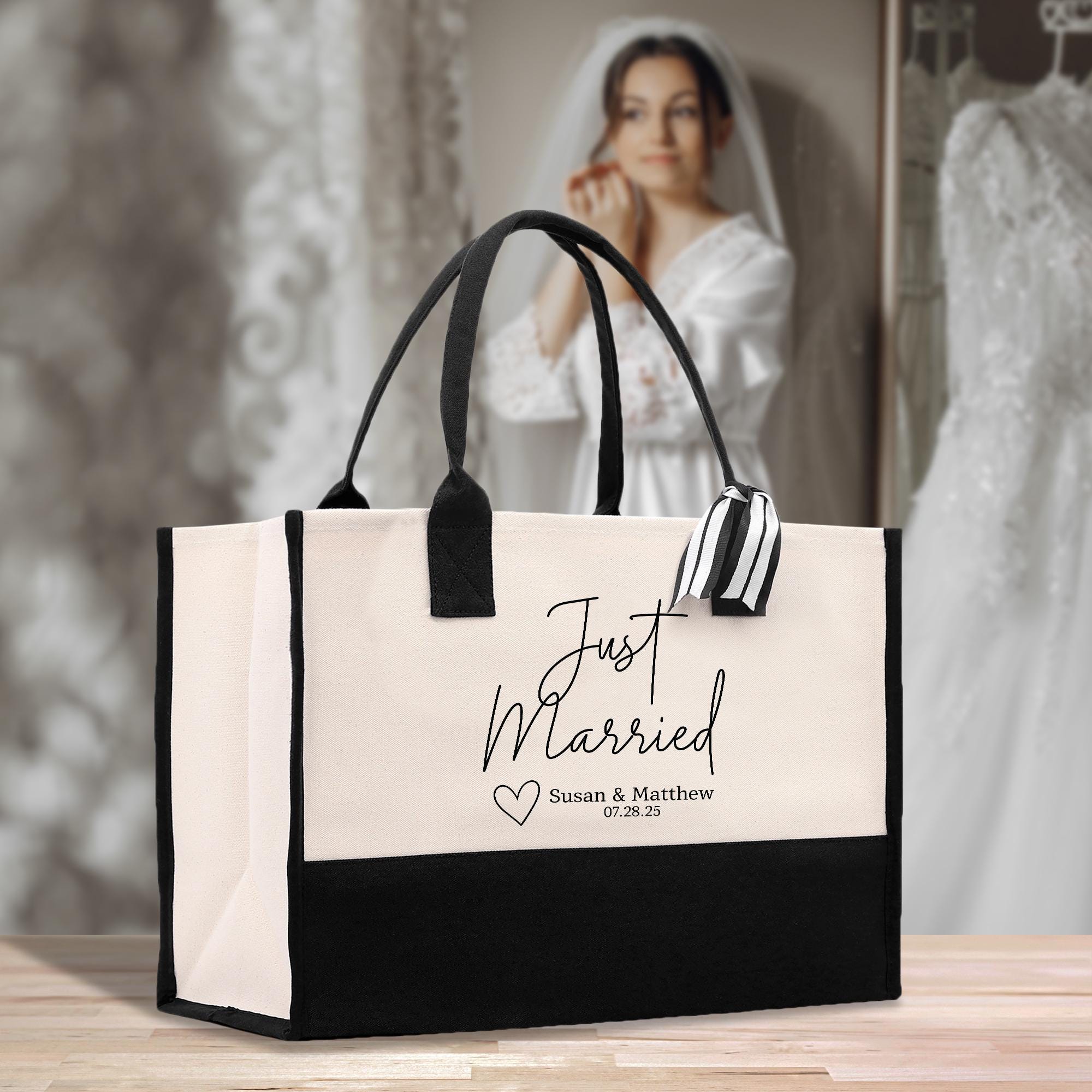 A white tote bag with the text &quot;Just Married Susan & Matthew 07:28:25&quot; printed on it, placed on a wooden surface. In the background, there is a blurred image of a woman wearing a white wedding dress.