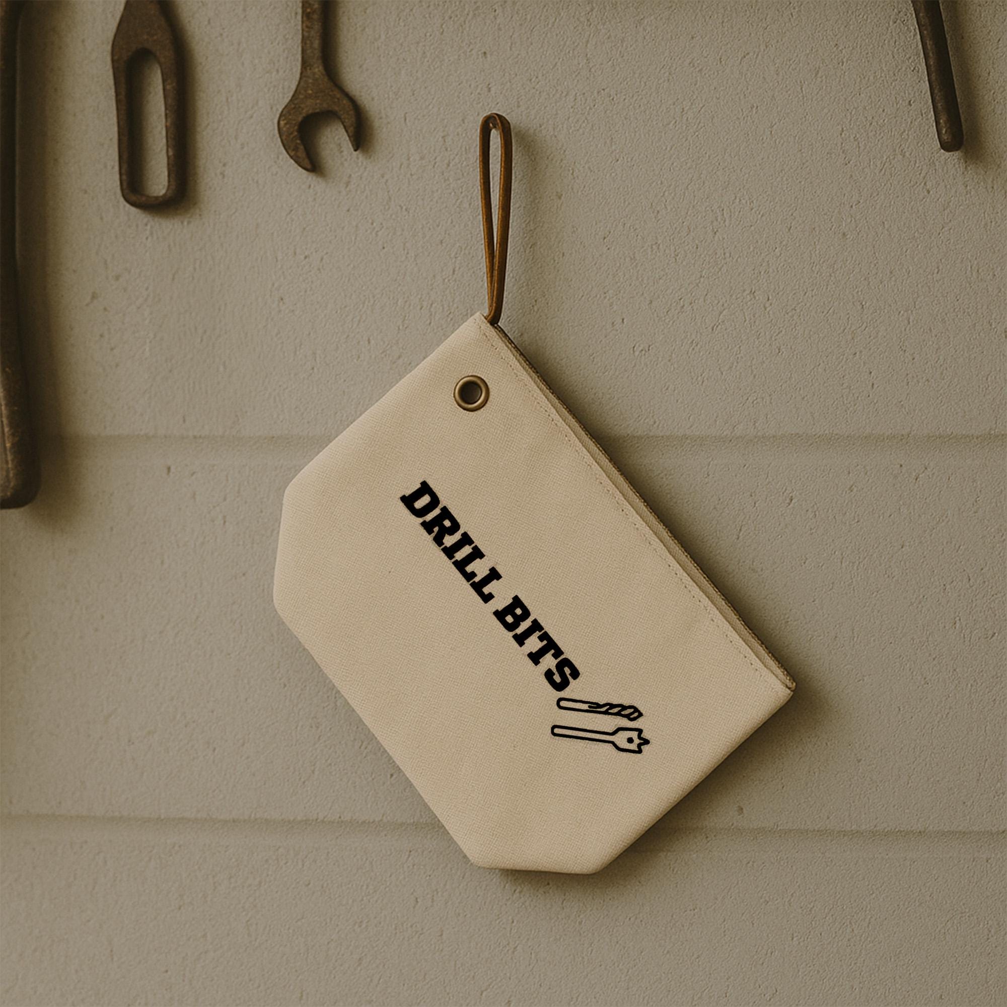 A beige canvas bag with the text &quot;DRILL Bits&quot; printed on it, hanging on a wall with various tools visible in the background.