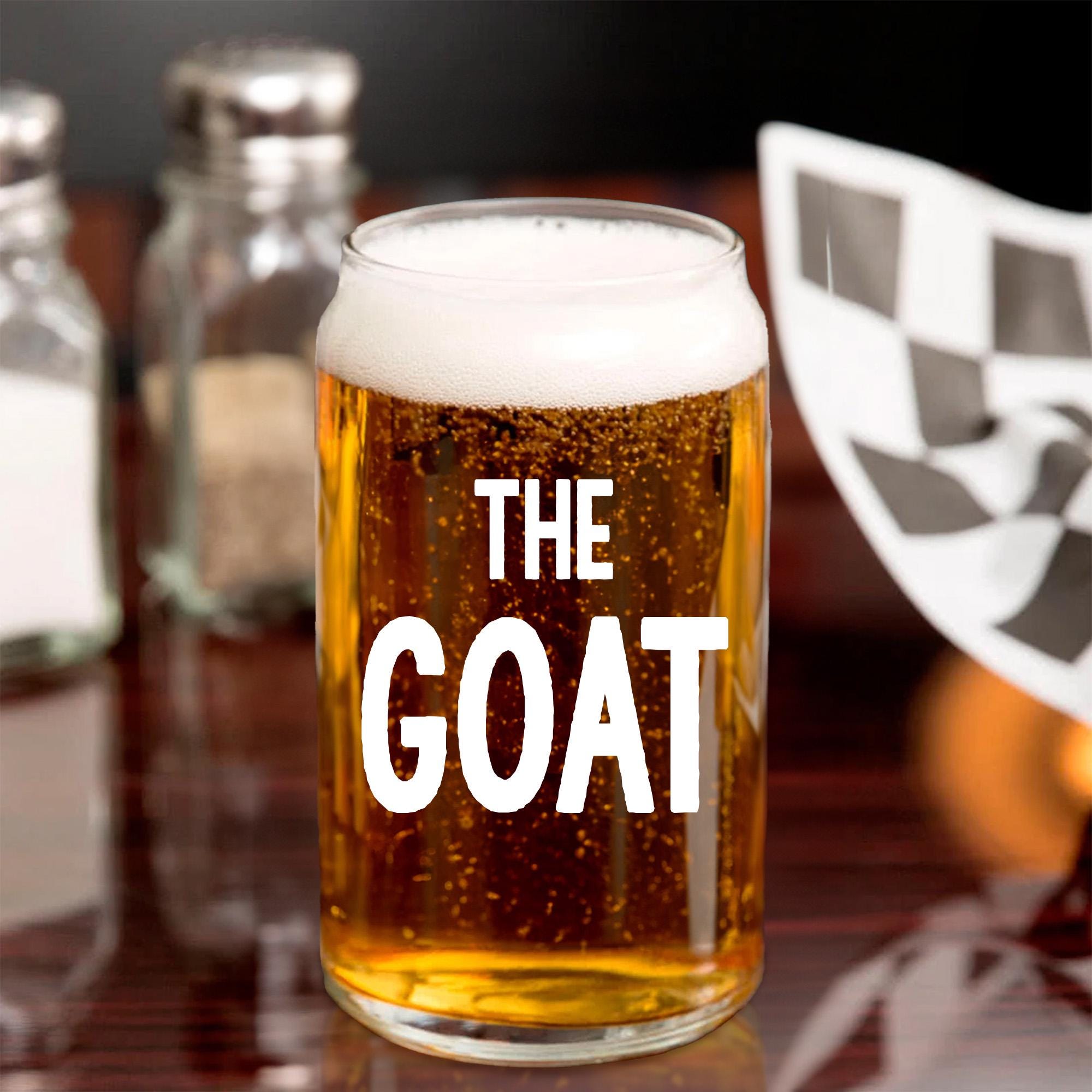 A glass of beer with the text &quot;THE GOAT&quot; printed on it, placed on a table with other items in the background.