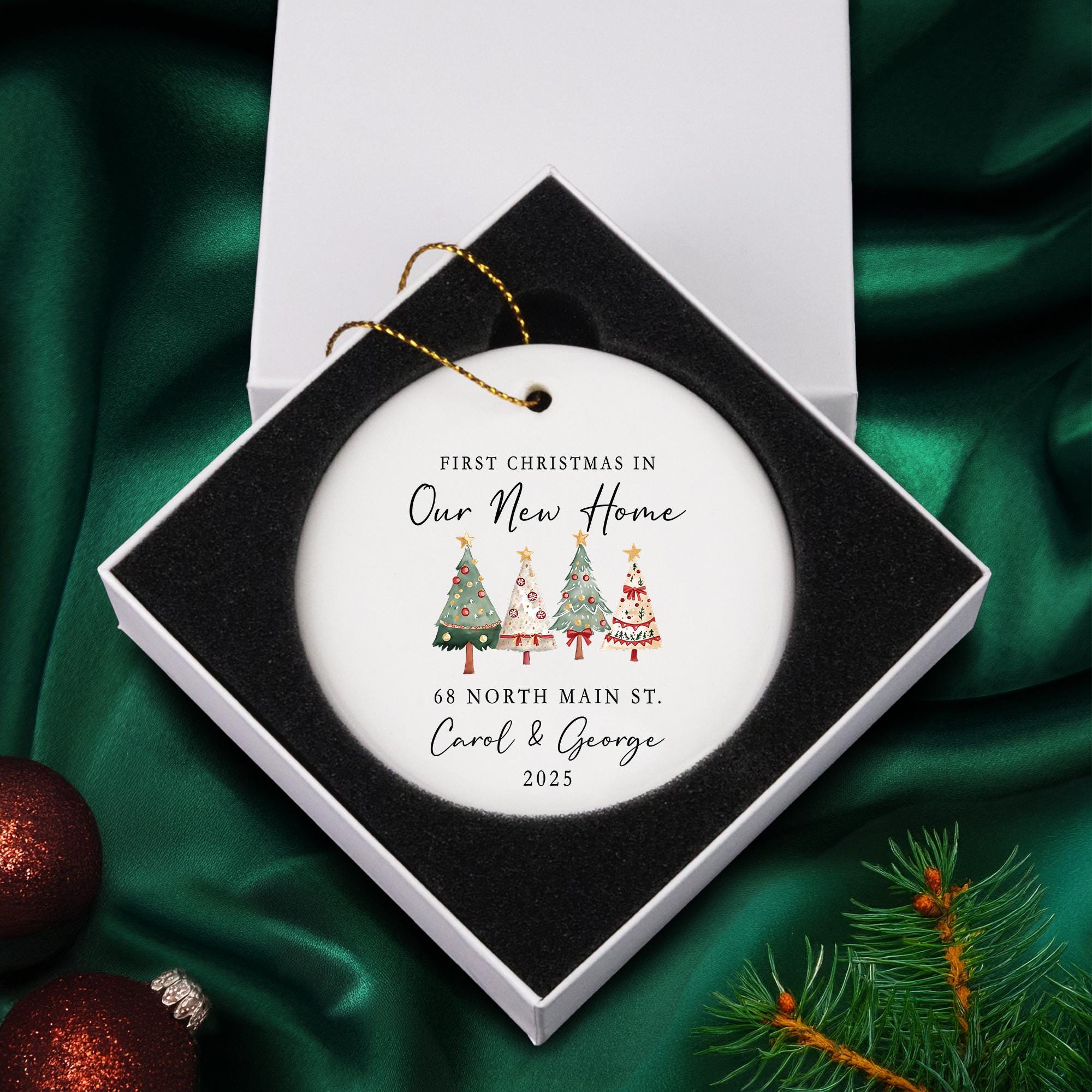 A white ornament with Christmas trees and text, placed in a white box on a green background.