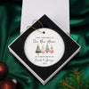A white ornament with Christmas trees and text, placed in a white box on a green background.