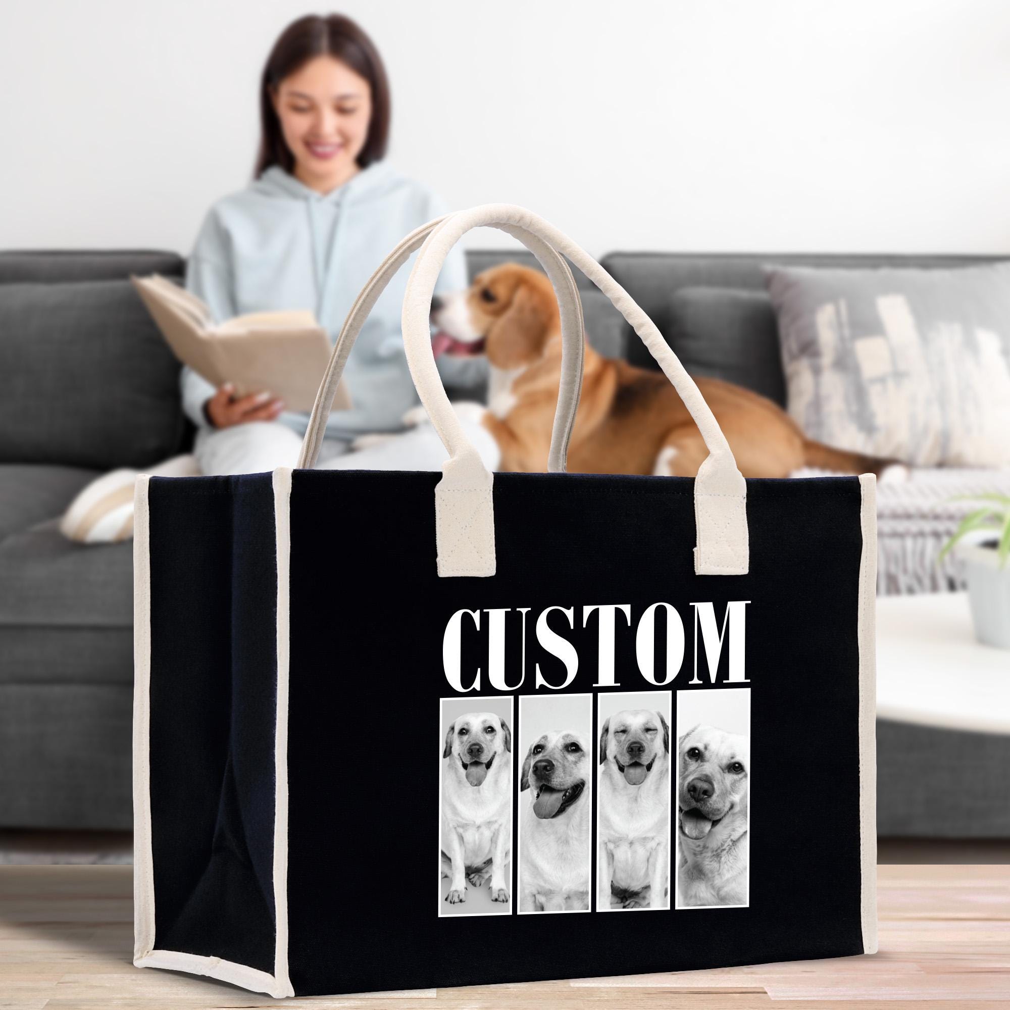 A woman is sitting on a couch, reading a book, while a dog is in the background. A black tote bag with a white handle and the word &quot;CUSTOM&quot; printed on it is placed in front of the couch.