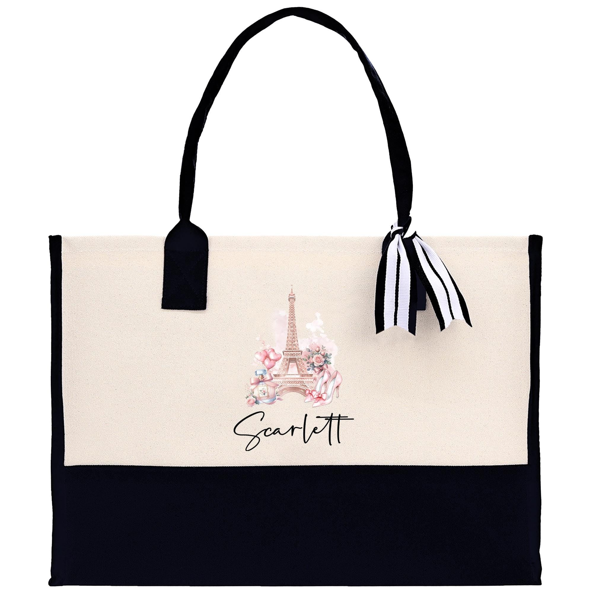 A beige tote bag with a black handle and a black and white striped ribbon attached to it. The bag has a printed image of the Eiffel Tower on it, and the name &quot;Scartlett&quot; is written below the image.