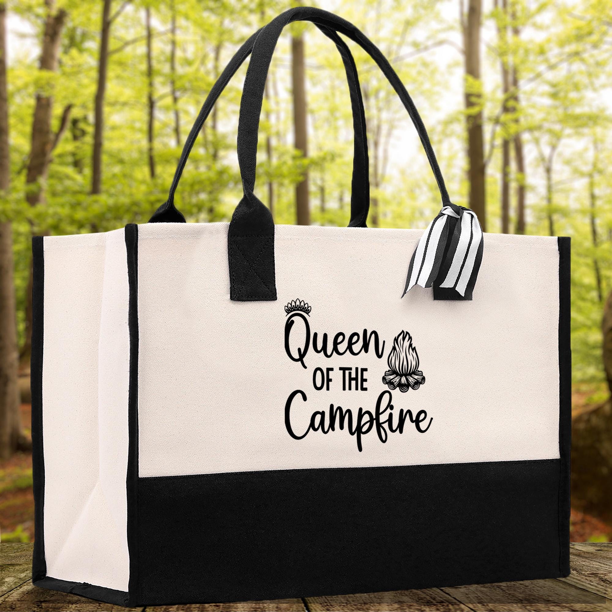 A black and white tote bag with the text "Queen of the Campfire" printed on it, placed on a wooden surface with a forested background.