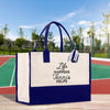 A white and blue tote bag with the text "Life Happens Tennis Helps" printed on it, placed on a wooden surface against a blurred background of a tennis court and trees.