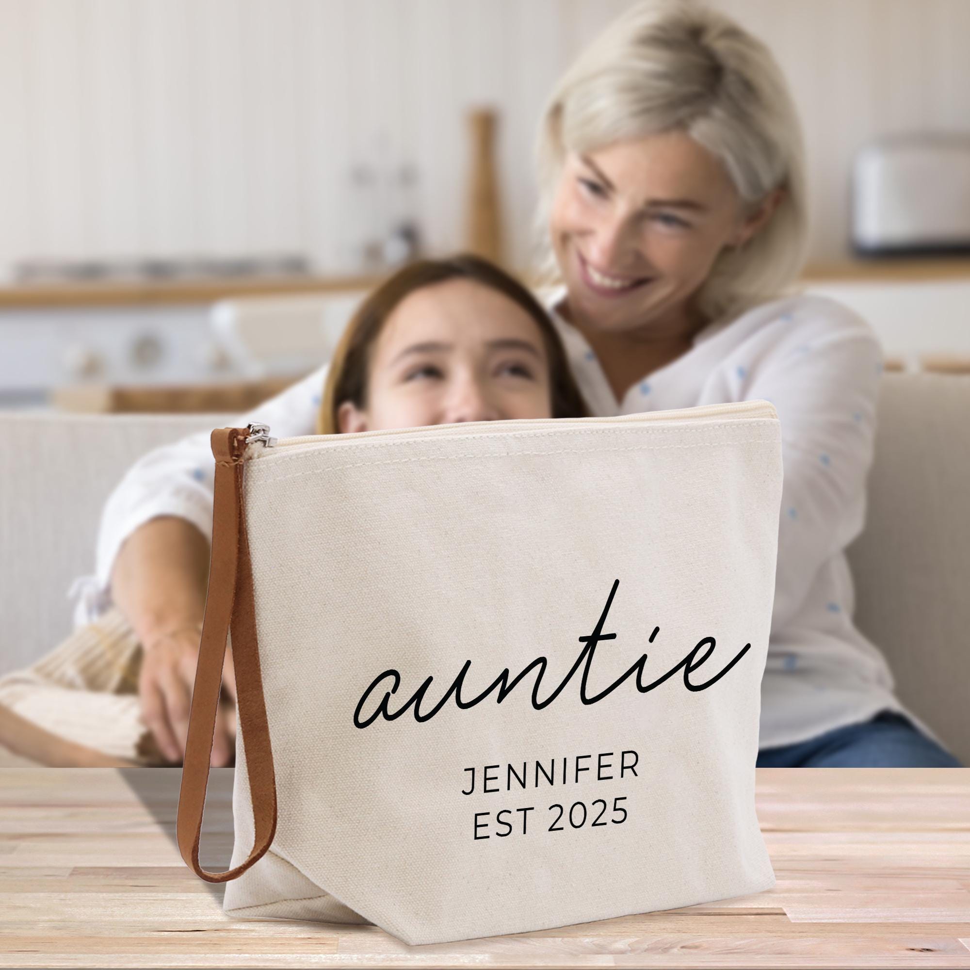 A woman is sitting on a couch, smiling, while a young girl is sitting behind her, peeking out from behind a beige tote bag with the word &quot;auntie&quot; printed on it.