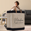 A beige tote bag with black trim and leather handles, featuring a personalized address printed on it. The bag is placed on a wooden surface, with a blurred figure of a woman in the background.