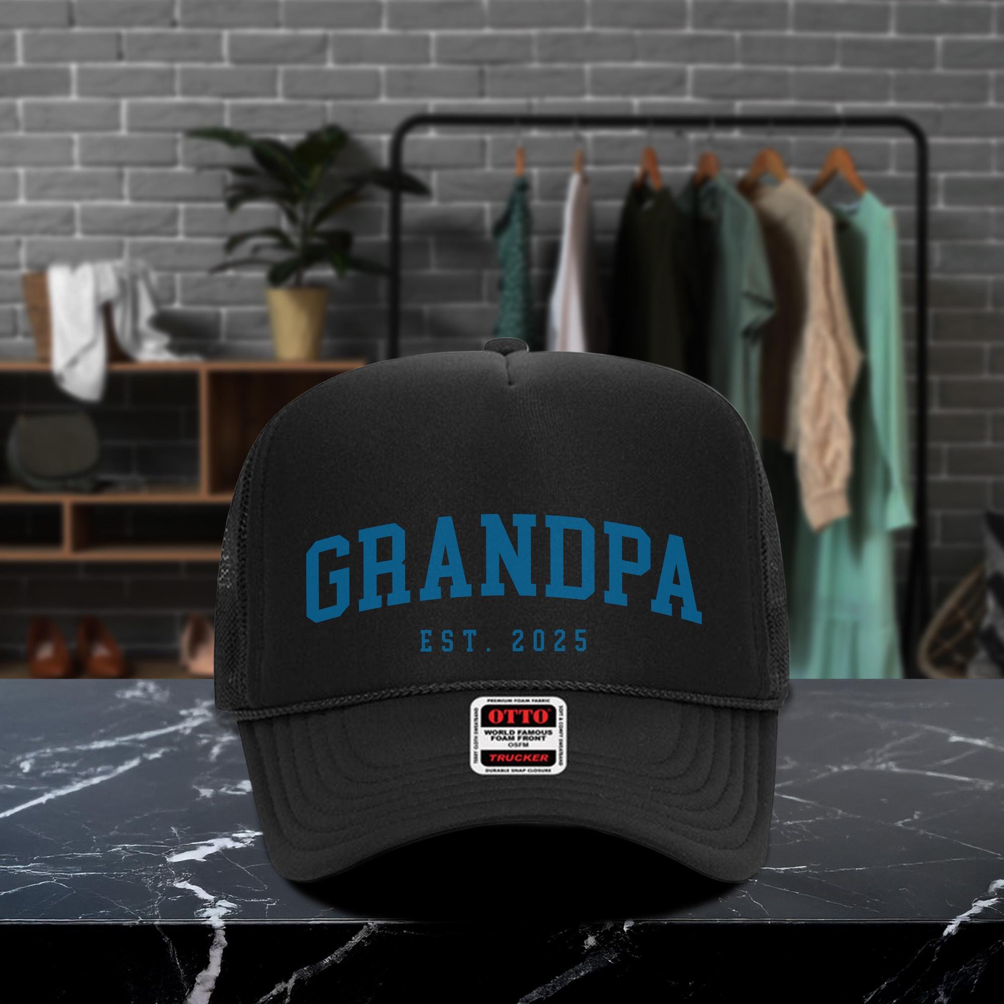 A black baseball cap with the text &quot;GRANDPA EST. 2025&quot; printed on it, placed on a marble countertop.