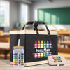 A black tote bag with a handle, a glass jar with a straw, and a small metal container with a label that says "Miss Magi" are displayed on a wooden surface.