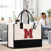 A large tote bag with a prominent red "M" on it, placed on a wooden surface in a room with a person sitting on a couch in the background.