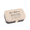 a rectangular business card holder with a tan and black color scheme. The holder has a gold clasp and displays the text "Olivia Williams REALTOR" and a contact number "123 456-7890" along with the website "Olivia@REALTOR.com".
