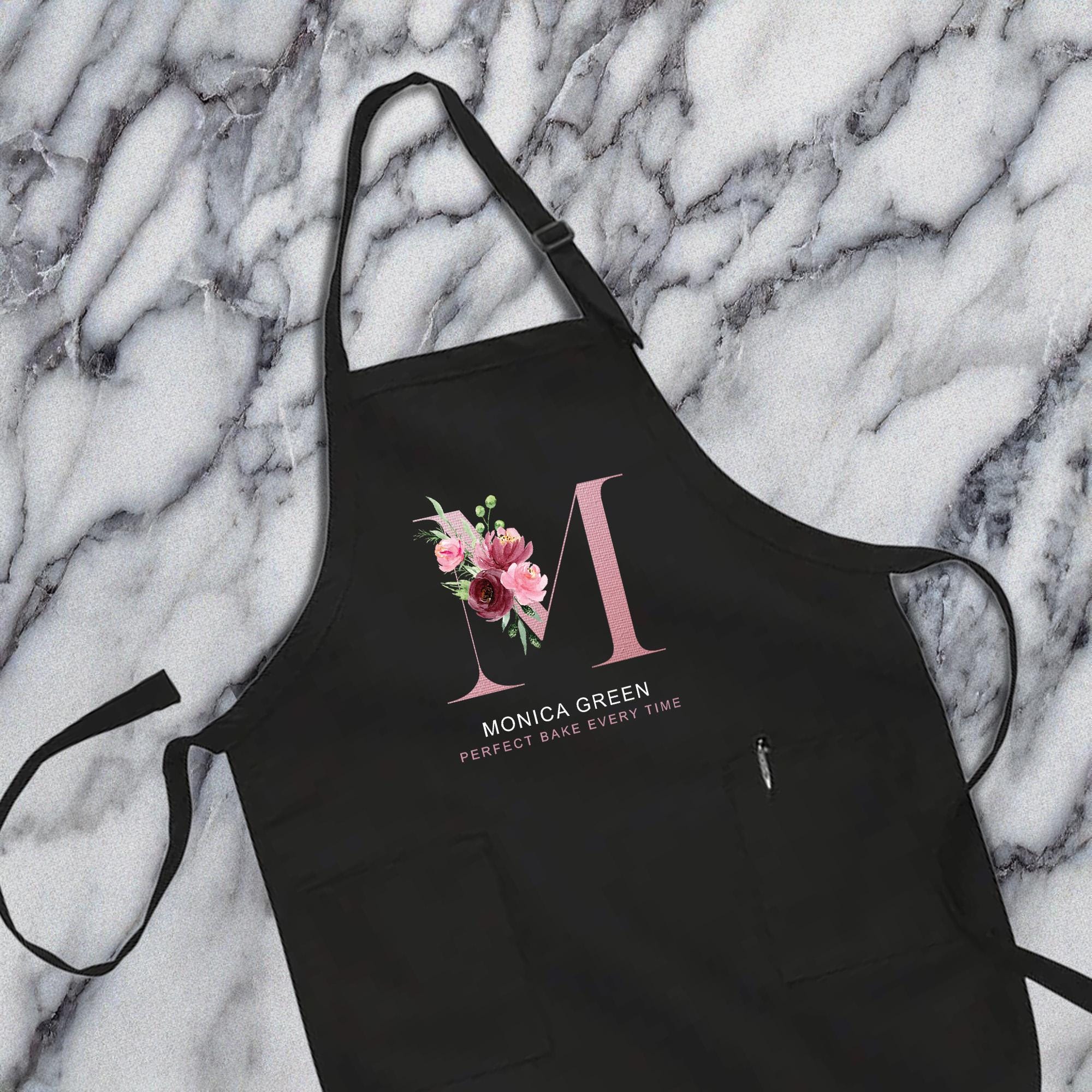A black apron with a pink floral design and the text &quot;MONICA GREEN&quot; printed on it, placed on a marble surface.