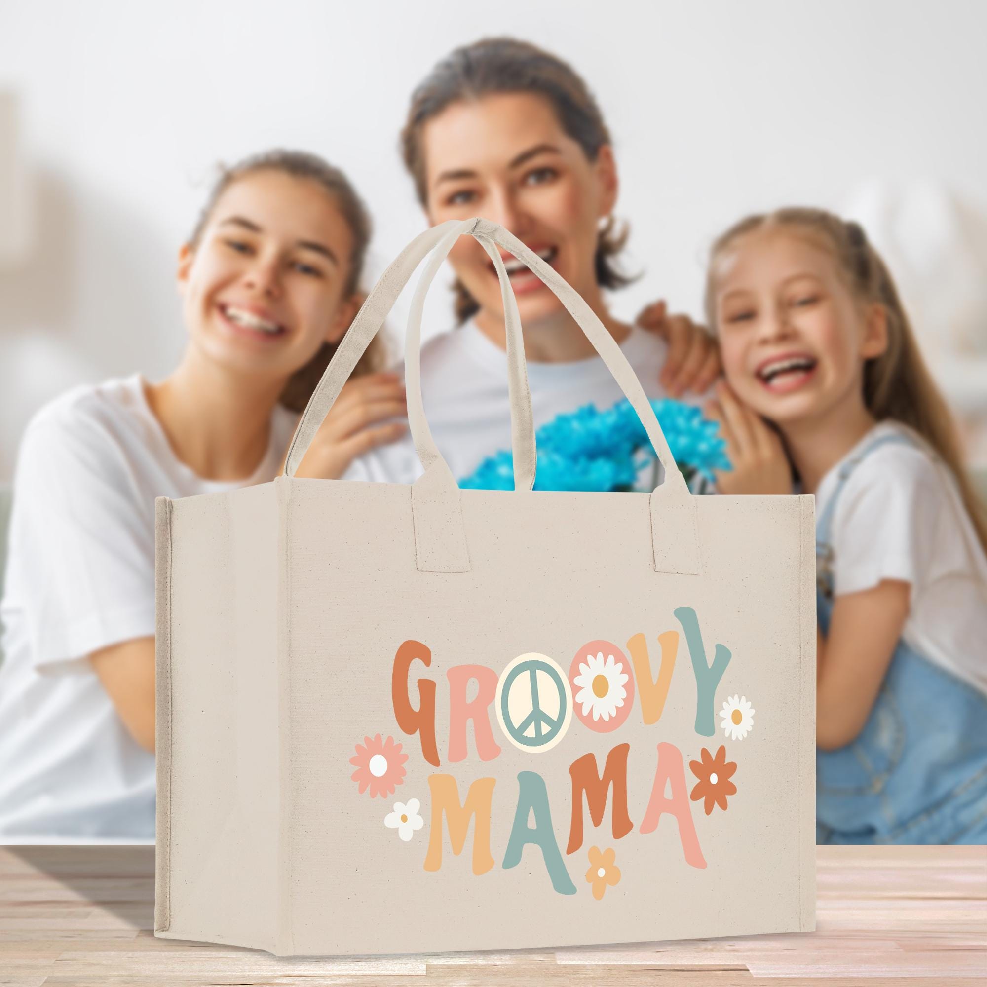 A beige tote bag with the text &quot;GROOVY MAMA&quot; printed on it, along with various colorful flowers and peace signs, is displayed in the foreground. In the background, there are two smiling women, one of whom is holding a bouquet of flowers.